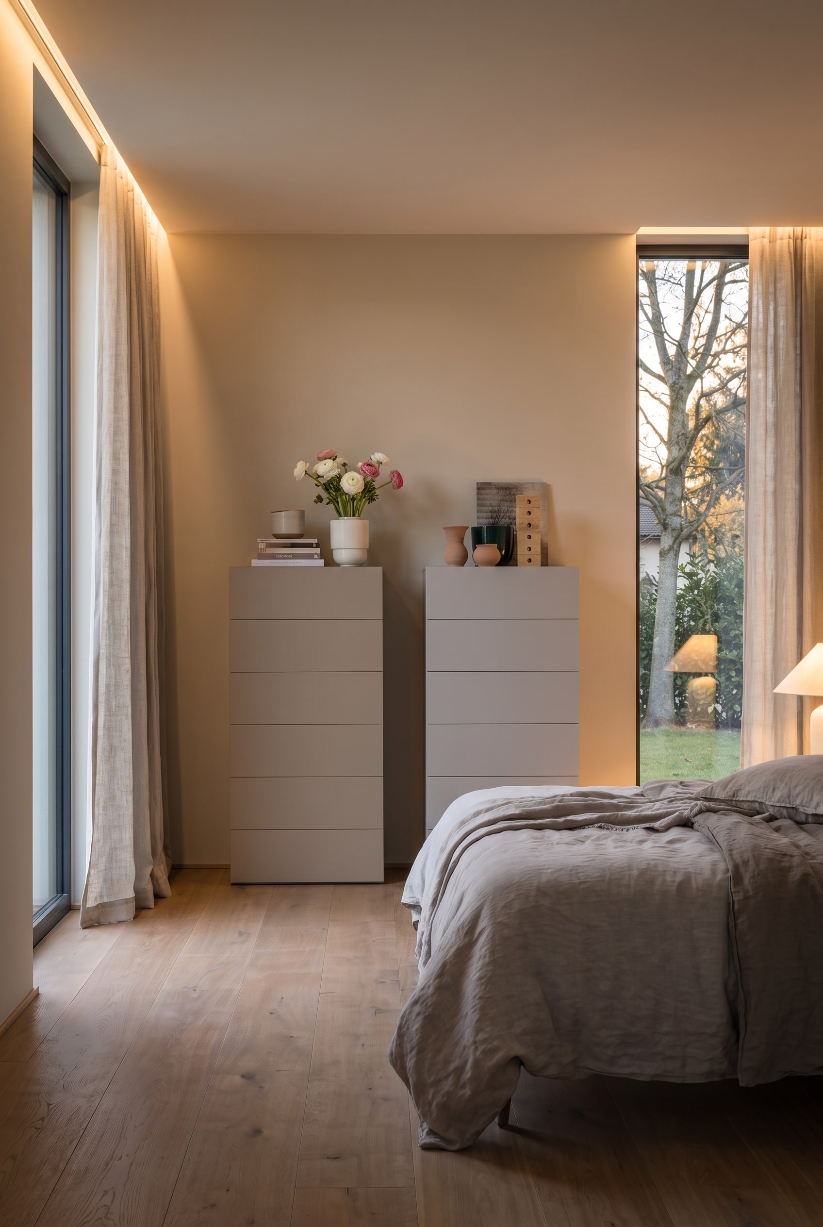 Zwei greige Hochkommoden in einem modernen Schlafzimmer mit Leinenbettw&auml;sche und bodentiefen Fenstern.