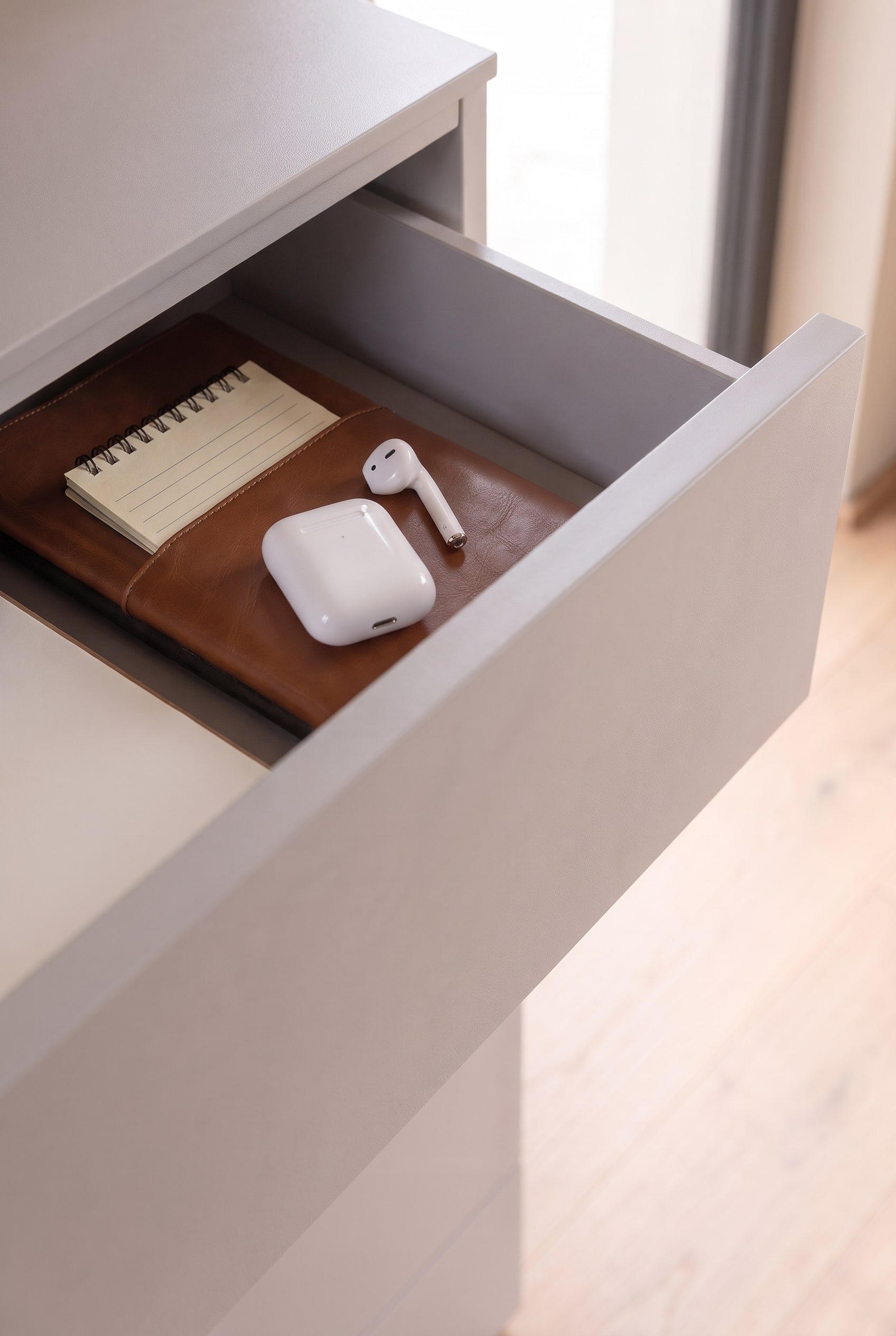 Geöffnete Schublade einer greigen Kommode mit einem Leder-Notizbuch und weißen AirPods.
